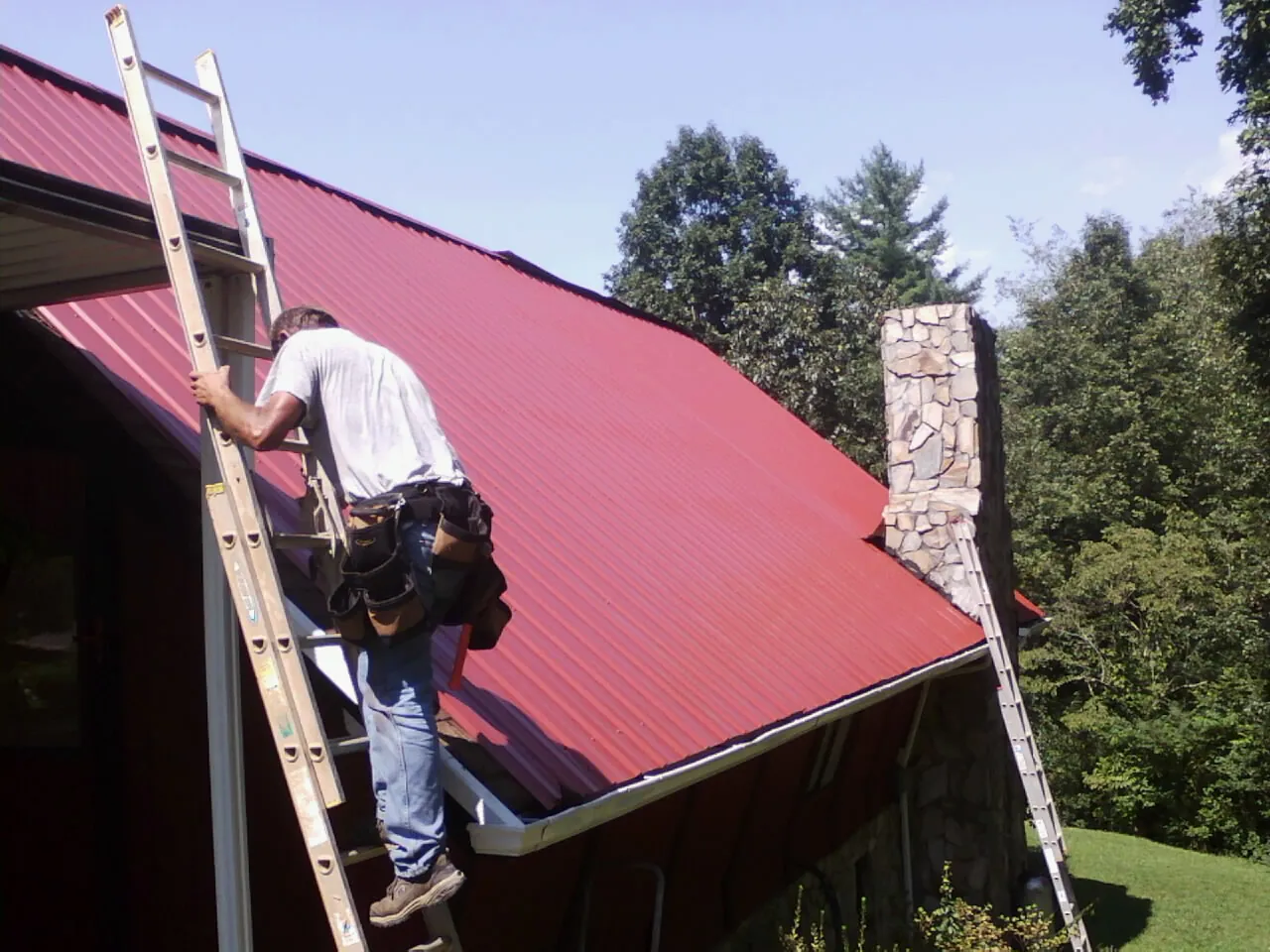 Roofer on a ladder beside a red metal roof and stone chimney — Soffit & Fascia Repair by Roofing Brothers in Bullard